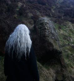 a man with long white hair standing in front of a large rock on the side of a hill