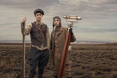 two men standing next to each other in the middle of an open field with a telescope