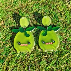 two green earrings sitting in the grass with faces drawn on them, one has an angry look