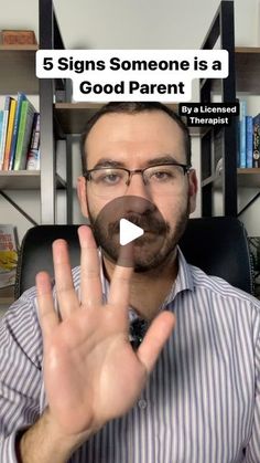 a man with glasses and a beard is making a hand gesture while sitting in front of a bookshelf