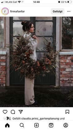 a woman standing in front of a brick building holding a wreath with pine cones on it