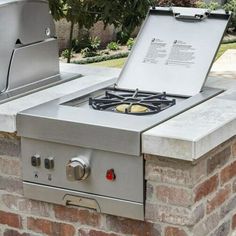 an outdoor grill with two burners on the top and one open lid, sitting next to a brick wall