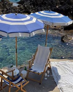 two beach chairs with blue and white umbrellas on the sand near an open pool