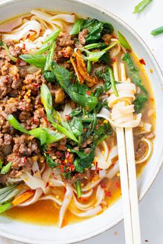 a white bowl filled with noodles, meat and veggies next to chopsticks