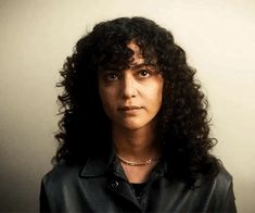 a woman with long curly hair wearing a black leather jacket and gold choker necklace