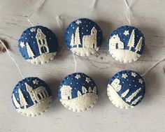 four blue and white ornaments hanging from strings on a wooden surface with snow covered buildings in the background