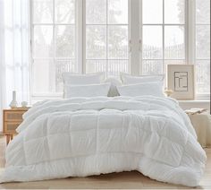 a bed with white comforter and pillows in front of two large windows on the floor