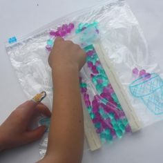 a person is drawing on some plastic bags with colored beads and scissors in it, while another hand holds a pencil