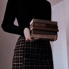 a woman is holding three books in her hands while wearing a black sweater and plaid skirt