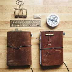 two brown notebooks sitting on top of a wooden table next to some metal clips