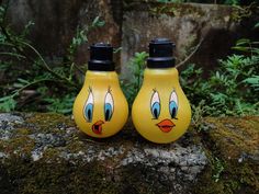 two yellow lightbulbs with faces painted on them sitting on a mossy rock