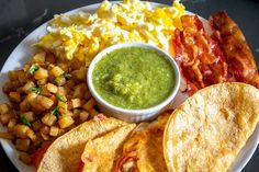 a white plate topped with tacos and bacon next to eggs, potatoes and guacamole