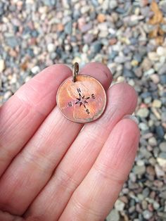 a hand holding a tiny compass charm on it's thumb, with gravel in the background