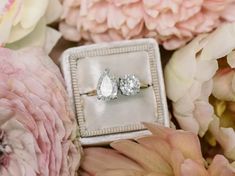 two diamond engagement rings sitting on top of a box surrounded by flowers and peonies