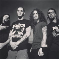 four men standing next to each other in front of a wall with tattoos on their arms