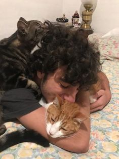 a man laying on top of a bed holding a cat next to his face with two other cats in the background