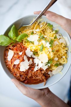 two hands holding a bowl of food with pasta, cheese and spinach on top