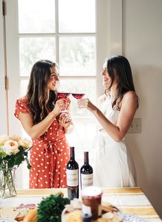 two women standing next to each other holding wine glasses in front of a window with flowers