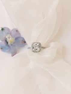 a diamond engagement ring sitting on top of a white cloth next to a blue flower