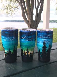 three painted tumbles sitting on top of a wooden table next to a tree and water