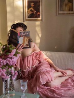 a woman sitting on a couch reading a book in front of a vase with flowers