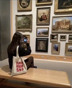 a woman sitting on a bench taking a photo with her cell phone in front of some paintings