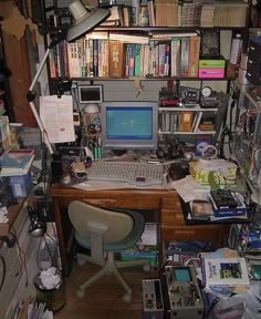 a cluttered desk with a computer and bookshelf in the background, along with many other office supplies