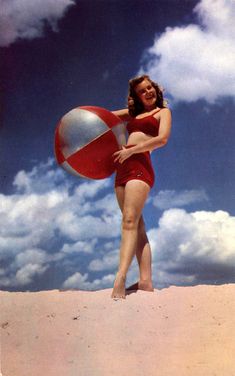 a woman in a red bathing suit holding a beach ball on the sand under a cloudy blue sky