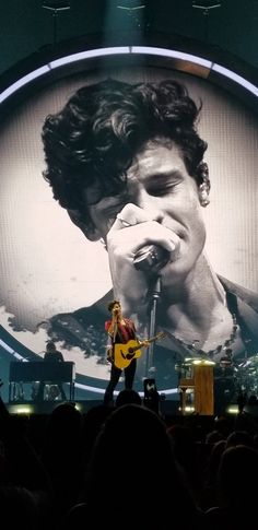 a man holding a guitar while standing on top of a stage next to a large screen