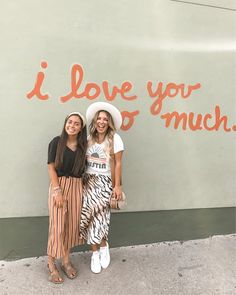 two women standing next to each other in front of a wall that says i love you much