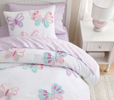 a white bed with pink and blue butterflies on it's comforter, next to a night stand