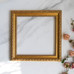 an empty gold frame sitting on top of a white marble counter next to flowers and greenery