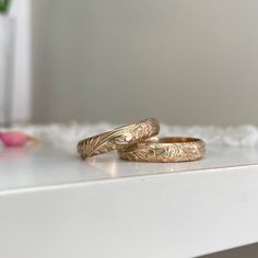 two gold wedding rings sitting on top of a white table next to a pink flower