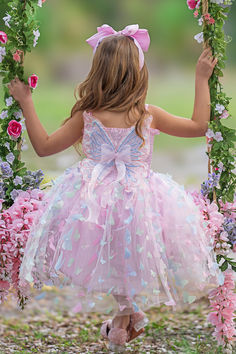 A little girl in a whimsical pink butterfly dress with delicate butterfly accents, standing on a floral swing adorned with flowers, exuding fairy-tale charm. Cute Multicolor Fairy Dress For Spring, Multicolor Fairy Style Dress-up Dresses, Fairy Style Multicolor Dress For Dress-up, Playful Sleeveless Spring Fairy Dress, Spring Sleeveless Playful Fairy Dress, Pink Sleeveless Dress With Butterfly Print, Cute Butterfly Dresses For Spring, Pink Fairy Princess Dress For Spring, Spring Multicolor Whimsical Princess Dress