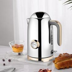 a silver and gold electric kettle on a table next to bread, jams and tea