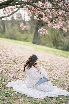 a mother holding her baby while sitting on a blanket under a tree with pink flowers