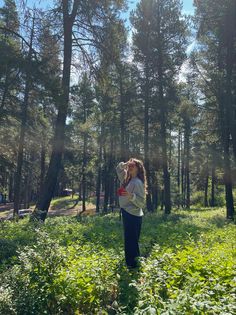 a woman standing in the middle of a forest drinking from a water bottle while looking up into the sky