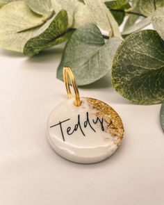 a white and gold pendant with the word teddy written on it next to some green leaves