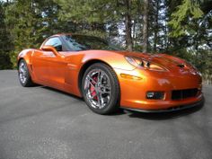 an orange sports car parked in a parking lot next to some trees and pine trees