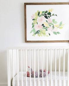 a white crib with pink and green flowers on the wall next to a framed painting