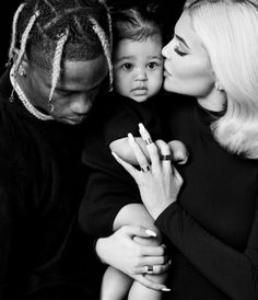 a black and white photo of a woman holding a baby while the man kisses her cheek