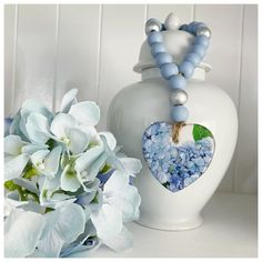 a white vase filled with blue flowers next to a heart shaped beaded necklace on a table