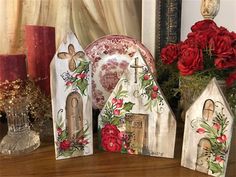 three painted wooden houses sitting on top of a table next to red roses and candles