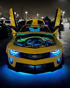 a yellow sports car with its hood open and lights on at night in a parking lot