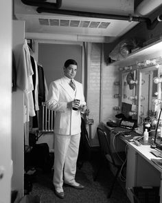 a man in a white suit and tie holding a cup standing in front of a mirror