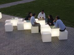 several people sitting on white cubes in the middle of a brick walkway at night