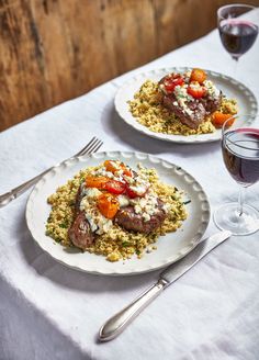 two white plates topped with meat and rice covered in sauce next to glasses of wine