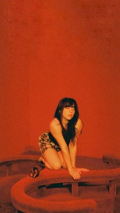 a woman sitting on top of a wooden bench in front of a red wall with her legs crossed