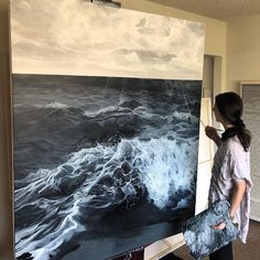 a woman standing in front of a large painting with waves on the water and clouds above her