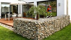 a large rock wall sitting in the grass next to a patio with chairs and an umbrella